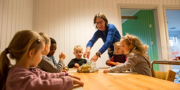 Barnehagelærer og seks barn som sitter rundt et bord. Illustrasjonsfoto