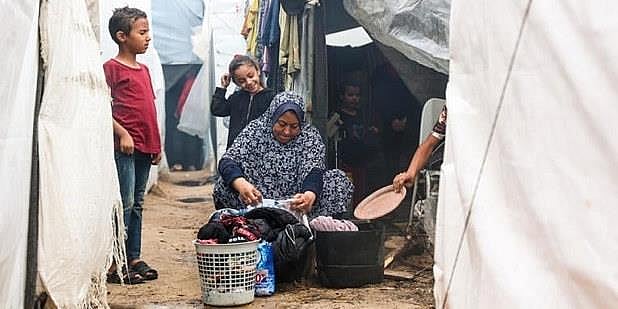 Foto fra en flyktningleir i Gaza, kvinne sitter utenfor et telt og ordner med klær, to barn ser på, sand, søle