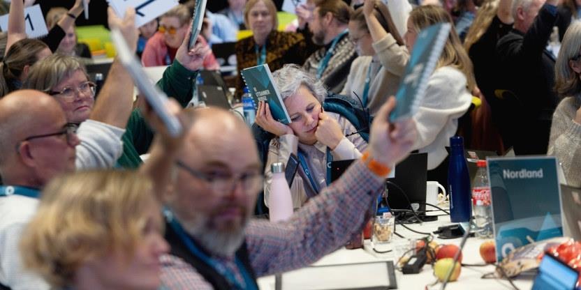 Bilde fra landsmøtesalen der delegater holder stemmekort i været.