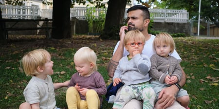 En barnehagelærer sitter på bakken, med to barn i fanget og to ved siden av seg.