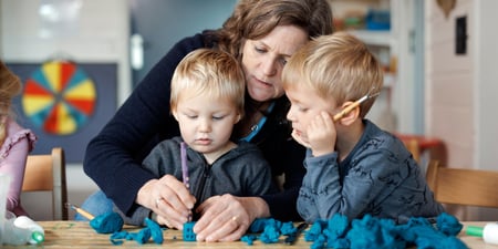 Barnehagelærer sitter med to barn ved bordet og leker med naturgummi. Inne