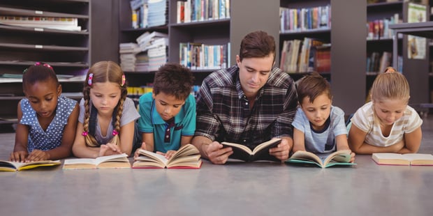 En voksen mann og fem barn ligger på gulvet og leser i hver sin bok i et bibliotek.