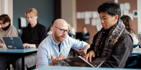 Mannlig lærer veileder elev ved pulten. Flere elever ved pultene rundt, videregående skole.