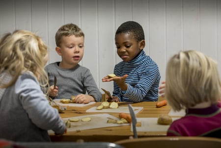 En mannlig barnehagelærer med minoritetsbakgrunn leker med to barnehagebarn i sandkassa. Det er blader fra trærne på bakken og sola skinner.