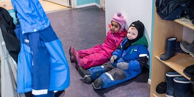 To blide barn sitter på gulvet i barnehagen. De har yttertøyet på. Foto.