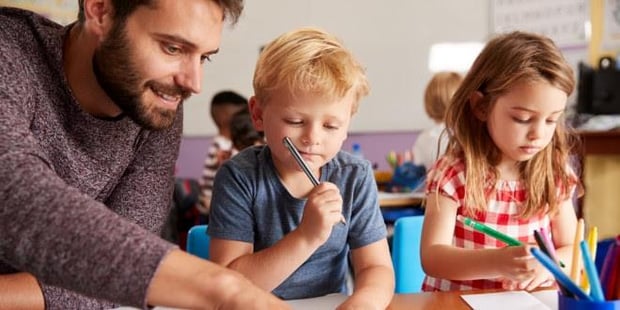 Smilende lærer so sitter ved pulten til to elever i småskolen og viser dem noe i bøkene. Illustrasjonsfoto