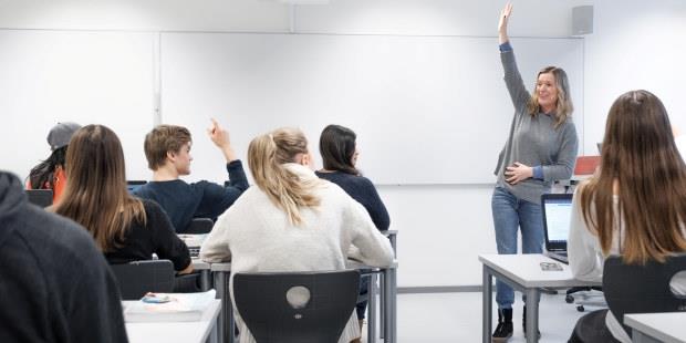 Bilde av lærer som rekker opp hånda og smiler til en klasse på videregående skole. 