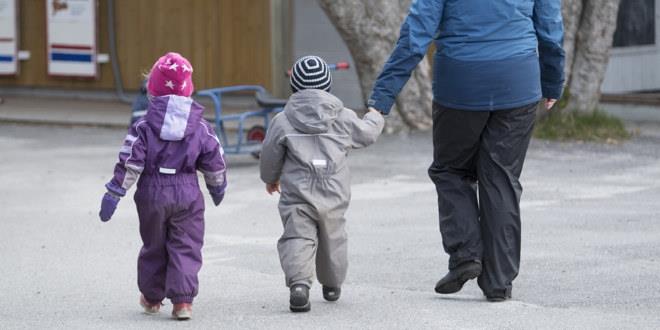 To små barn i en barnehage går mot barnehagebygget med ryggen mot kameraet. Det ene barnet leier en voksen.