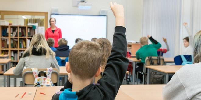 Bilde fra et klasserom. Vi ser lærer i front og en elev som rekker opp hånda sett bakfra. 