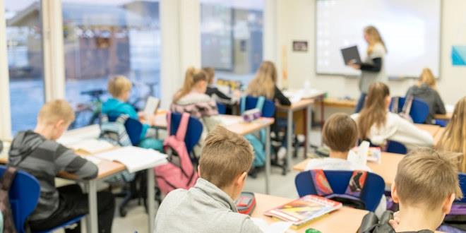 foto fra et fullt klasserom der elevene ses bakfra, ved tavla foran ses en kvinnelig lærer 