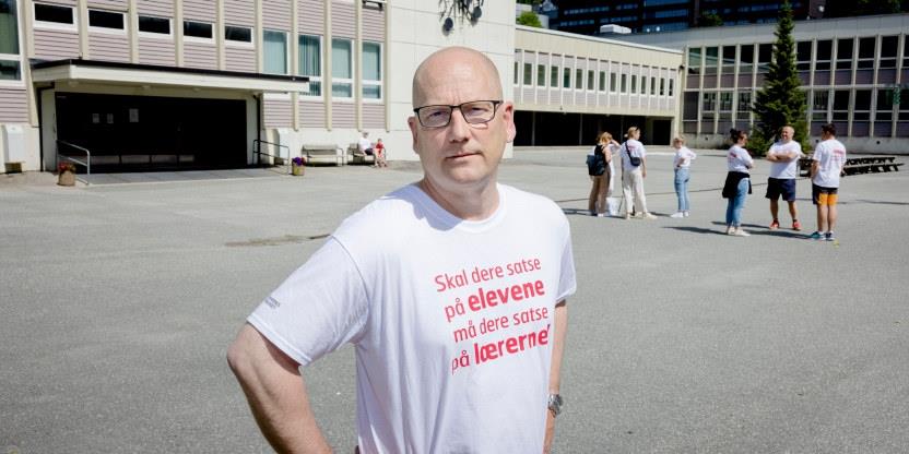Steffen Handal med hvit streike t-skjorte med rød skrift «Skal dere satse på elevene, må dere satse på lærerne» i en skolegård med hendene på hofta og seriøst blikk. 