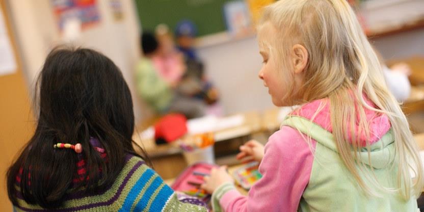 To barn sitter på skolepulten sin med ryggen til. Den ene har lyst hår og den andre har mørkt hår.