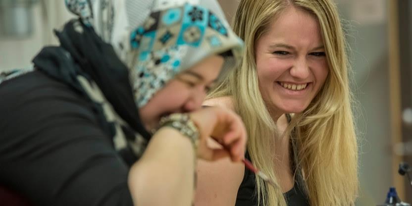 Nærbilde av to videregående elever som ler. Hun fremste har på seg et fargerikt sjal på hodet og holder hånden foran munnen. Hun bakerste har langt lyst hår og gliser. 