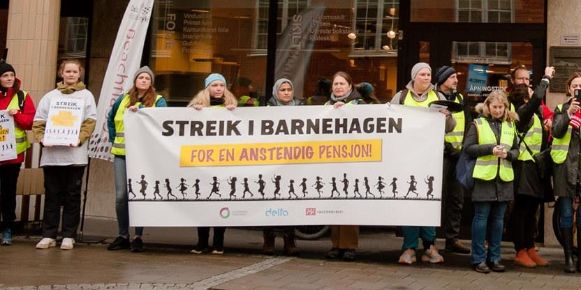 Utsnitt med rundt 20 streikende barnehagelærere som står ute og holder et stort banner der det står at man streiker. 