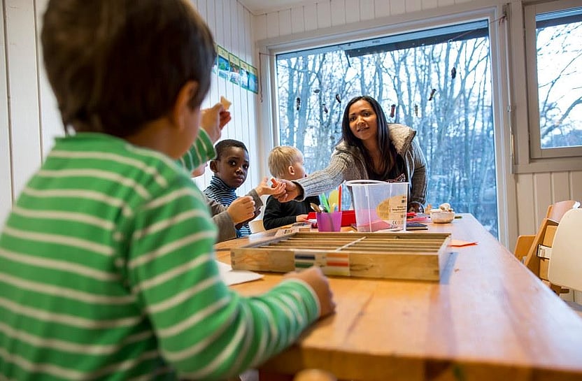 Barn sitter rundt et bord i en barnehage. En barnehaglærer sitter ved bord-enden og gir noe til et av barna.