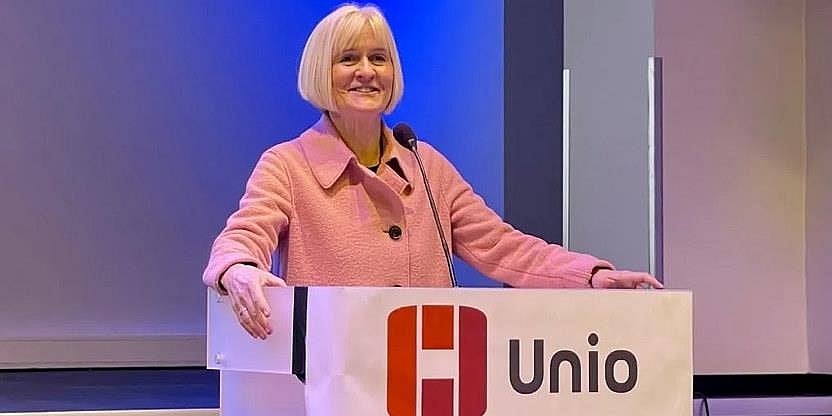 Ragnhild Lied står foran en talerstol på Unios konferanse. Hun har kort hvitt hår med pannelugg og har på seg en rosa dressjakke. Hun holder talestoler med begge hender og ser utover forsamlingen og smiler. 