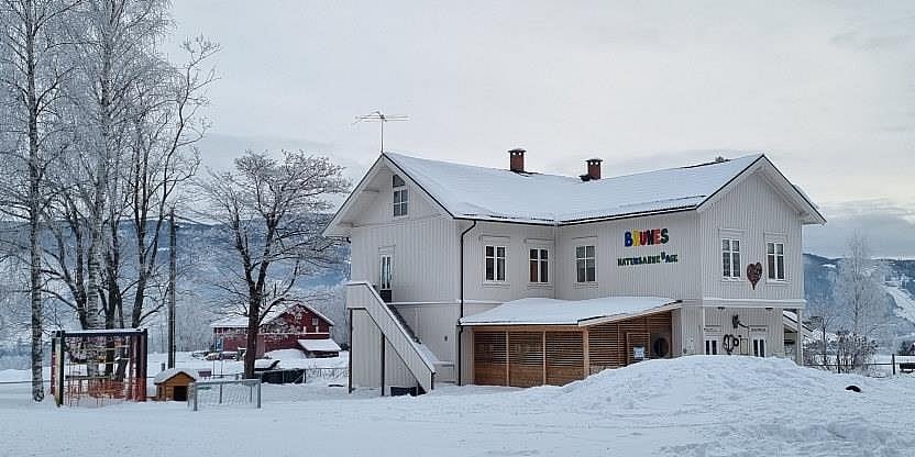 Hvitmalt barnehage, tatt utendørs, med mye snø, fjell i bakgrunnen, og noen trær og en lekeplass. 