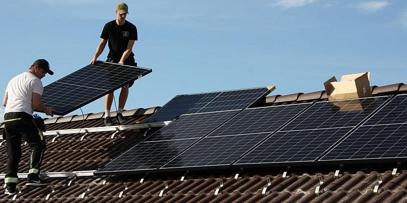 To arbeidsfolk løfter på plass et solcellepanel på taket på en bygning som allerede har flere solcellepaneler på plass.