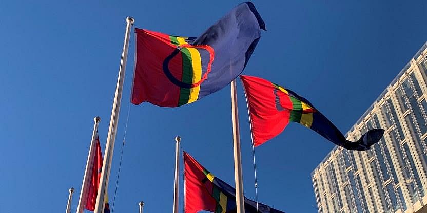 Samiske flagg blafrer i vinden. Det er sol og i bakgrunnen skimtes et moderne bygg i glass og stål.